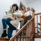 Un prieten care ajută la căratul unor cutii pe scări, într-o zi de mutare, zâmbind