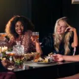 Grup de prieteni care râd la o masă, seara, într-o atmosferă relaxată, alături de un cuplu